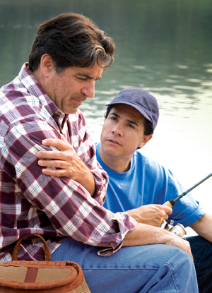 Homme assis près d'un lac avec une canne à pêche, se tenant le bras et ayant l'air bouleversé. Homme plus jeune assis à côté de lui, ayant l'air inquiet.